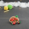 sur un fond gris foncé, il y a un panier crocheté en topaze garni de fruits rouges miniatures : fraises, cerises et baies variées (framboises, mûres,...). Certains fruits sont disposés à l'avant du panier. À l'arrière plan, sur la gauche il y a 2 pelotes jaune et verte et un crochet. En fond est disposé un nuage de ouate et une guirlande lumineuse.