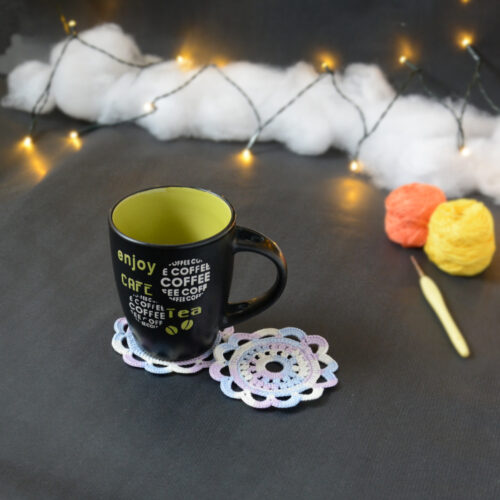 Une tasse noire à l'intérieur vert clair est posée sur deux sous-verres ronds crochetés dans un dégradé parme et lilas. En fond, à droite, deux pelotes de coton (jaune et orange) et un crochet. En arrière-plan, des guirlandes lumineuses sont disposées sur du rembourrage blanc façon nuages.