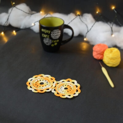 Deux sous-verres ronds crochetés dans un dégradé jaune et orange sont posés sur un fond gris sombre. En fond, à droite, deux pelotes de coton (jaune et orange) et un crochet. Sur la gauche, il y a une tasse noire à l'intérieur vert clair. En arrière-plan, des guirlandes lumineuses sont disposées sur du rembourrage blanc façon nuages.