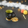 Une tasse noire à l'intérieur vert clair est posée sur deux sous-verres ronds crochetés dans un dégradé jaune et orange. En fond, à droite, deux pelotes de coton (jaune et orange) et un crochet. En arrière-plan, des guirlandes lumineuses sont disposées sur du rembourrage blanc façon nuages.