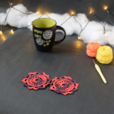 Deux sous-verres ronds crochetés en rouge et aubergine sont posés sur un fond gris sombre. En fond, à droite, deux pelotes de coton (jaune et orange) et un crochet. Sur la gauche, il y a une tasse noire à l'intérieur vert clair. En arrière-plan, des guirlandes lumineuses sont disposées sur du rembourrage blanc façon nuages.