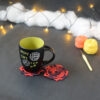 Une tasse noire à l'intérieur vert clair est posée sur deux sous-verres ronds crochetés en rouge et aubergine. En fond, à droite, deux pelotes de coton (jaune et orange) et un crochet. En arrière-plan, des guirlandes lumineuses sont disposées sur du rembourrage blanc façon nuages.
