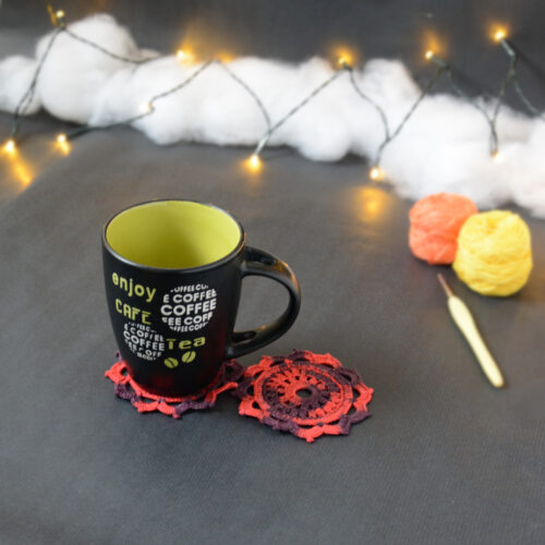 Une tasse noire à l'intérieur vert clair est posée sur deux sous-verres ronds crochetés en rouge et aubergine. En fond, à droite, deux pelotes de coton (jaune et orange) et un crochet. En arrière-plan, des guirlandes lumineuses sont disposées sur du rembourrage blanc façon nuages.