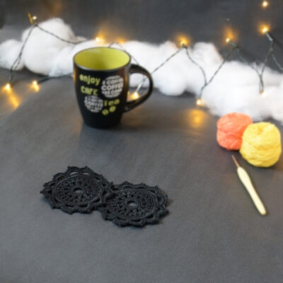 Deux sous-verres ronds crochetés en noir sont posés sur un fond gris sombre. En fond, à droite, deux pelotes de coton (jaune et orange) et un crochet. Sur la gauche, il y a une tasse noire à l'intérieur vert clair. En arrière-plan, des guirlandes lumineuses sont disposées sur du rembourrage blanc façon nuages.