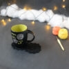 Une tasse noire à l'intérieur vert clair est posée sur deux sous-verres ronds crochetés en noir. En fond, à droite, deux pelotes de coton (jaune et orange) et un crochet. En arrière-plan, des guirlandes lumineuses sont disposées sur du rembourrage blanc façon nuages.