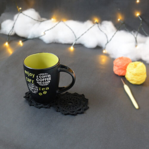 Une tasse noire à l'intérieur vert clair est posée sur deux sous-verres ronds crochetés en noir. En fond, à droite, deux pelotes de coton (jaune et orange) et un crochet. En arrière-plan, des guirlandes lumineuses sont disposées sur du rembourrage blanc façon nuages.