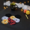 Huit sous-verres ronds crochetés sont posés sur un fond gris sombre. Ils sont de différentes couleurs (dégradé parme et lilas, dégradé orange et jaune, dégradé violet et aubergine et uni noir. En fond, à droite, deux pelotes de coton (jaune et orange) et un crochet. Sur la gauche, il y a une tasse noire à l'intérieur vert clair. En arrière-plan, des guirlandes lumineuses sont disposées sur du rembourrage blanc façon nuages.
