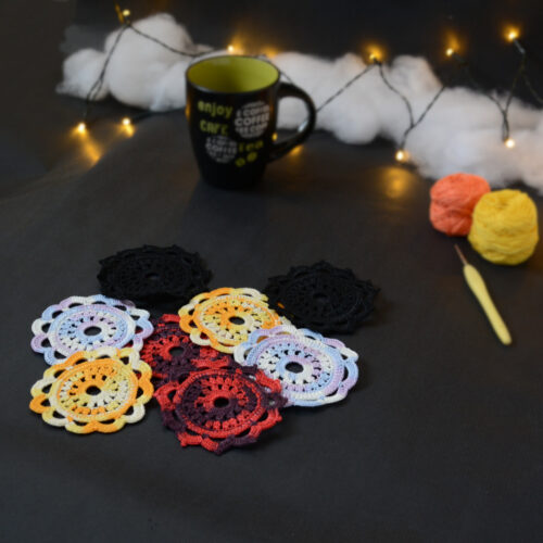 Huit sous-verres ronds crochetés sont posés sur un fond gris sombre. Ils sont de différentes couleurs (dégradé parme et lilas, dégradé orange et jaune, dégradé violet et aubergine et uni noir. En fond, à droite, deux pelotes de coton (jaune et orange) et un crochet. Sur la gauche, il y a une tasse noire à l'intérieur vert clair. En arrière-plan, des guirlandes lumineuses sont disposées sur du rembourrage blanc façon nuages.