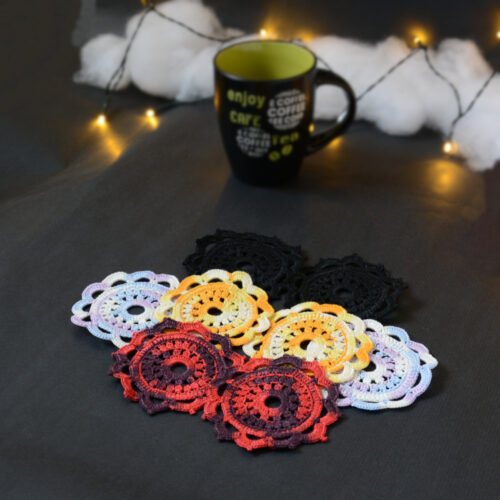 Huit sous-verres ronds crochetés sont posés sur un fond gris sombre. Ils sont de différentes couleurs (dégradé parme et lilas, dégradé orange et jaune, dégradé violet et aubergine et uni noir. En fond il y a une tasse noire à l'intérieur vert clair et des guirlandes lumineuses sont disposées sur du rembourrage blanc façon nuages.