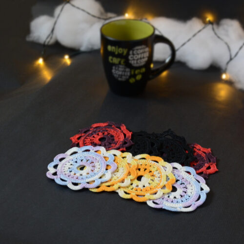 Huit sous-verres ronds crochetés sont posés sur un fond gris sombre. Ils sont de différentes couleurs (dégradé parme et lilas, dégradé orange et jaune, dégradé violet et aubergine et uni noir. En fond il y a une tasse noire à l'intérieur vert clair et des guirlandes lumineuses sont disposées sur du rembourrage blanc façon nuages.