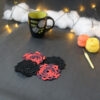 Quatre sous-verres ronds crochetés sont posés sur un fond gris sombre, 2 sont noirs et les 2 autres sont rouge et aubergine. Ils sont disposés en alternance de couleurs et en carré. En fond, à droite, deux pelotes de coton (jaune et orange) et un crochet. Sur la gauche, il y a une tasse noire à l'intérieur vert clair. En arrière-plan, des guirlandes lumineuses sont disposées sur du rembourrage blanc façon nuages.