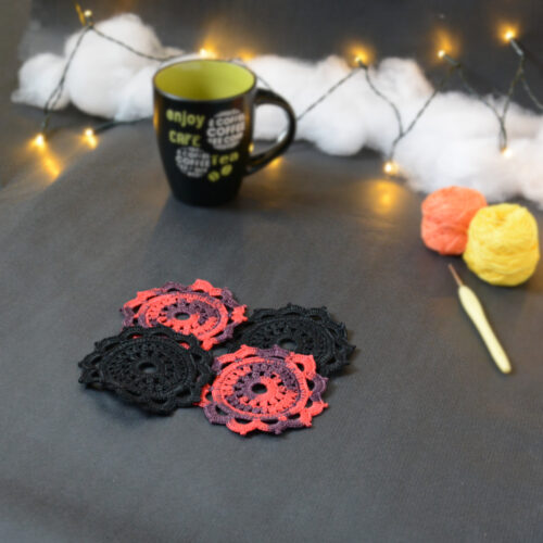 Quatre sous-verres ronds crochetés sont posés sur un fond gris sombre, 2 sont noirs et les 2 autres sont rouge et aubergine. Ils sont disposés en alternance de couleurs et en carré. En fond, à droite, deux pelotes de coton (jaune et orange) et un crochet. Sur la gauche, il y a une tasse noire à l'intérieur vert clair. En arrière-plan, des guirlandes lumineuses sont disposées sur du rembourrage blanc façon nuages.