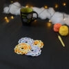 Quatre sous-verres ronds crochetés sont posés sur un fond gris sombre, 2 sont en dégradé orange et jaune et les 2 autres sont en dégradé parme et lilas. Ils sont disposés en alternance de couleurs et en carré. En fond, à droite, deux pelotes de coton (jaune et orange) et un crochet. Sur la gauche, il y a une tasse noire à l'intérieur vert clair. En arrière-plan, des guirlandes lumineuses sont disposées sur du rembourrage blanc façon nuages.