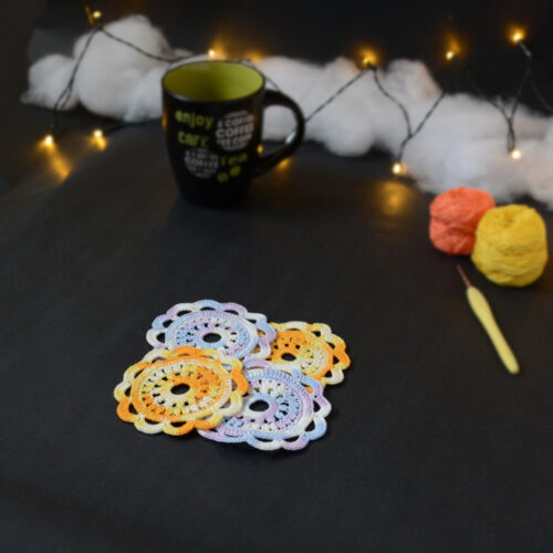 Quatre sous-verres ronds crochetés sont posés sur un fond gris sombre, 2 sont en dégradé orange et jaune et les 2 autres sont en dégradé parme et lilas. Ils sont disposés en alternance de couleurs et en carré. En fond, à droite, deux pelotes de coton (jaune et orange) et un crochet. Sur la gauche, il y a une tasse noire à l'intérieur vert clair. En arrière-plan, des guirlandes lumineuses sont disposées sur du rembourrage blanc façon nuages.