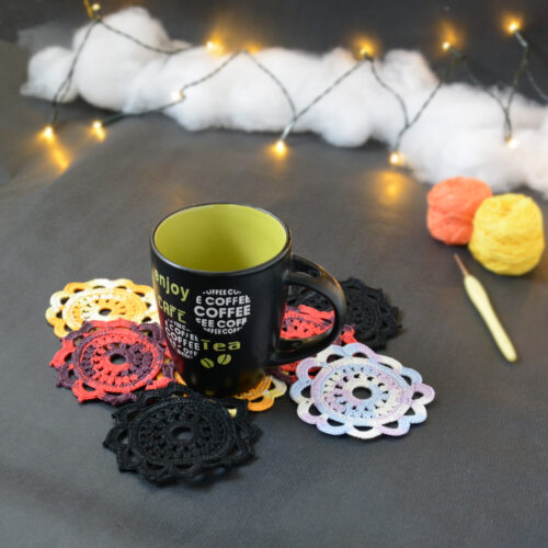 Huit sous-verres ronds crochetés sont posés sur un fond gris sombre. Ils sont de différentes couleurs (dégradé parme et lilas, dégradé orange et jaune, dégradé violet et aubergine et uni noir. Une tasse noire à l'intérieur vert clair est posée sur les sous-verres. En fond, à droite, deux pelotes de coton (jaune et orange) et un crochet. En arrière-plan, des guirlandes lumineuses sont disposées sur du rembourrage blanc façon nuages.