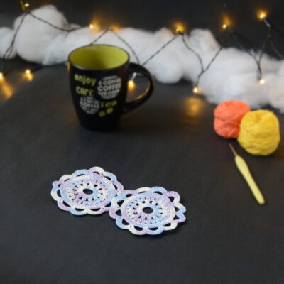 Deux sous-verres ronds crochetés dans un dégradé parme et lilas sont posés sur un fond gris sombre. En fond, à droite, deux pelotes de coton (jaune et orange) et un crochet. Sur la gauche, il y a une tasse noire à l'intérieur vert clair. En arrière-plan, des guirlandes lumineuses sont disposées sur du rembourrage blanc façon nuages.