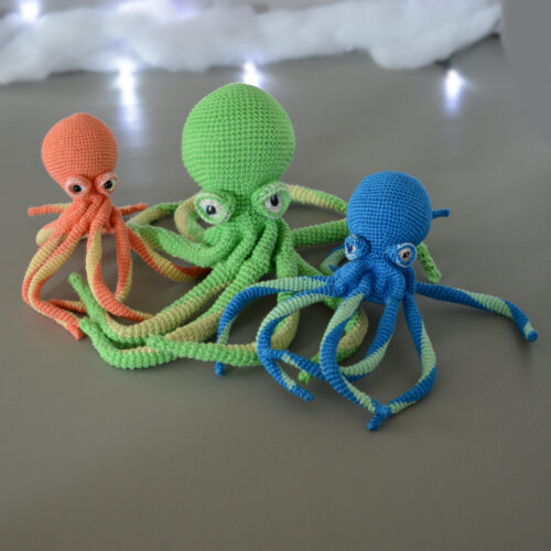 Un trio de poulpe au crochet est posé face à nous. Ils sont posés les uns sur les autres comme liés par leurs tentacules. Ils sont sur un fond gris avec en arrière-plan une guirlande lumineuse ainsi q'un nuage de ouate. Le poulpe le plus à gauche est de couleur corail et jaune pastel, celui du milieu est plus grand que les 2 autres et est vert et vert anis, enfin le dernier est bleu électrique et vert d'eau.
