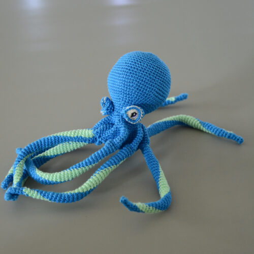 Gros plan sur un poulpe crocheté en bleu électrique et vert d'eau. Il est posé légèrement orienté vers la gauche sur un fond gris.