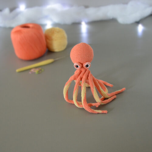Un poulpe crocheté en corail et jaune pastel, dressé sur ses tentacules, est posé sur un fond gris avec en fond 2 pelotes de la même couleur que le poulpe, un crochet et des anneaux marqueurs. En arrière plan, il y a une guirlande lumineuse et un nuage de ouate.