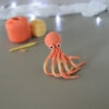 Un poulpe crocheté en corail et jaune pastel, dressé sur ses tentacules, est posé sur un fond gris avec en fond 2 pelotes de la même couleur que le poulpe, un crochet et des anneaux marqueurs. En arrière plan, il y a une guirlande lumineuse et un nuage de ouate.