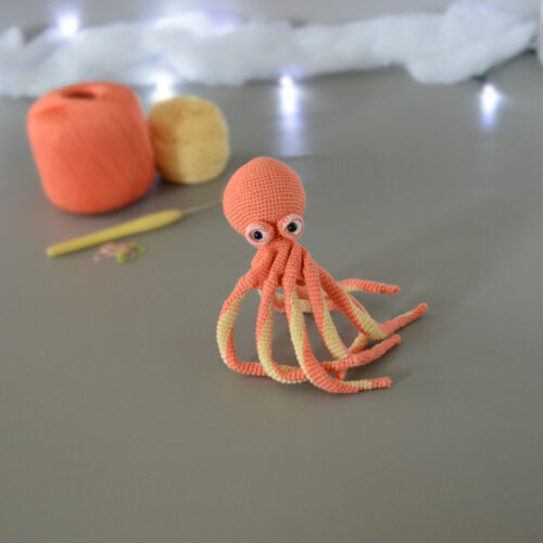 Un poulpe crocheté en corail et jaune pastel, dressé sur ses tentacules, est posé sur un fond gris avec en fond 2 pelotes de la même couleur que le poulpe, un crochet et des anneaux marqueurs. En arrière plan, il y a une guirlande lumineuse et un nuage de ouate.
