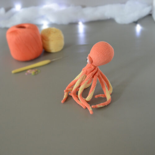 Un poulpe crocheté en corail et jaune pastel, dressé sur ses tentacules, est posé, tourné vers la gauche sur un fond gris avec en fond 2 pelotes de la même couleur que le poulpe, un crochet et des anneaux marqueurs. En arrière plan, il y a une guirlande lumineuse et un nuage de ouate.