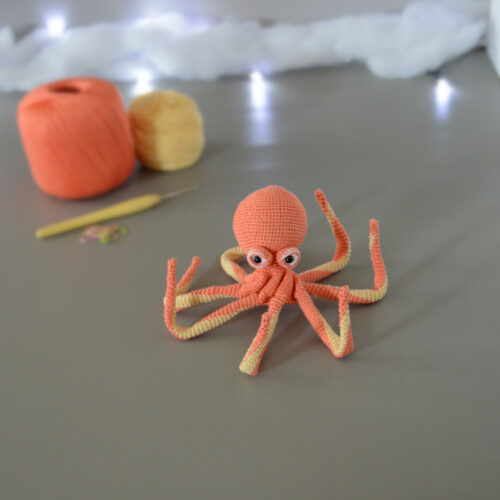 Un poulpe crocheté en corail et jaune pastel est posé avec les tentacules relevées sur un fond gris avec en fond 2 pelotes de la même couleur que le poulpe, un crochet et des anneaux marqueurs. En arrière plan, il y a une guirlande lumineuse et un nuage de ouate.