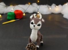 Amigurumi représentant un cerf brun et crème, derrière une pomme de pin, avec des bois crochetés couleur crème. En arrière-plan il y a des pelotes rouge et verte, un crochet ainsi qu'une guirlande lumineuse et des nuages de ouate.