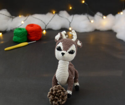 Amigurumi représentant un cerf brun et crème, derrière une pomme de pin, avec des bois crochetés couleur crème. En arrière-plan il y a des pelotes rouge et verte, un crochet ainsi qu'une guirlande lumineuse et des nuages de ouate.