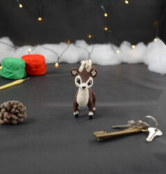 Amigurumi représentant un petit cerf brun et crème avec des bois crochetés couleur crème. Sur la gauche se trouve une pomme de pin. En arrière-plan il y a des pelotes rouge et verte, un crochet ainsi qu'une guirlande lumineuse et des nuages de ouate. Un trousseau de clés est posé au premier plan.