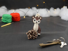 Amigurumi représentant un petit cerf brun et crème, derrière une pomme de pin, avec des bois crochetés couleur crème. En arrière-plan il y a des pelotes rouge et verte, un crochet ainsi qu'une guirlande lumineuse et des nuages de ouate. Un trousseau de clés est posé au premier plan.