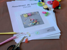 Des pages du patron de l'assortiment de bonbons sont posés sur un fond orangé. Dans le coin supérieur droit de ces pages, se trouve différents bonbons (frites, bananes, grosse fraise, réglisses, anneau apollo, bouteille de cola, etc). Dans le coin inférieur gauche sont disposés plusieurs crochets ainsi que des anneaux marqueurs et une paire de ciseaux. À l'arrière-plan on distingue une guirlande lumineuse.