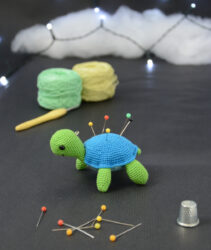 Amigurumi représentant une tortue verte et bleue crochetée, tournée vers la gauche. Elle a des épingles piquées dans sa carapace. Elle est posée sur un fond sombre avec en fond, des pelotes de coton vert et jaune ainsi qu’un crochet. En arrière plan, il y a une guirlande lumineuse et un nuage de ouate. À l'avant-plan sont disposés d'autres épingles ainsi qu'un dés à coudre.