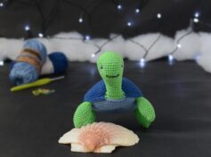 Amigurumi représentant une tortue verte à carapace bleue, vue de face, posée sur un coquillage, sur un fond noir avec des pelotes bleues, un crochet et une guirlande lumineuse en arrière-plan.