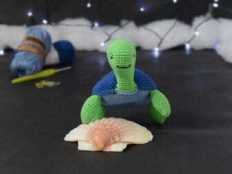 Géraldine la tortue Catona 78 Amigurumi représentant une tortue verte à carapace bleue, vue de face, posée sur un coquillage, sur un fond noir avec des pelotes bleues, un crochet et une guirlande lumineuse en arrière-plan.