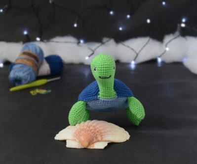 Amigurumi représentant une tortue verte à carapace bleue, vue de face, posée sur un coquillage, sur un fond noir avec des pelotes bleues, un crochet et une guirlande lumineuse en arrière-plan.