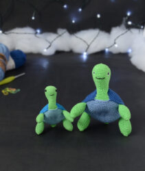 Deux tortues crochetées identiques en modèle mais de tailles différentes, posées côte à côte, dressées sur leurs pattes arrières et orientées face à l’objectif. La plus petite se trouve à gauche, la plus grande à droite. En arrière-plan flou : deux pelotes de fil bleu, un crochet jaune et quelques anneaux marqueurs, le tout posé sur un fond sombre avec une guirlande lumineuse.