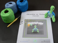 Sur un fond foncé est posé une page du patron de Géraldine la tortue. Sur le côté droit de cette page se trouve la tortue crochetée en vert et bleu, dressée sur ses pattes arrières. Dans le coin supérieur gauche de la page il y a trois pelotes de coton vert, bleu et bleu clair avec un crochet jaune.