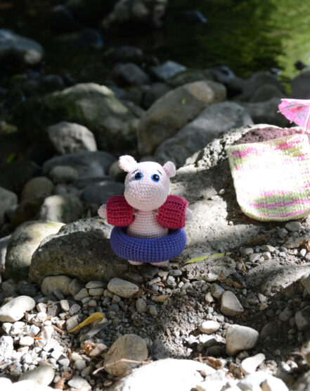 un hippopotame crocheté est posé sur des pierres en bord de rivière que l'on aperçoit contrebas. Il est rose pâle avec de grands yeux bleus. Il a une grosse bouée violette au tour de la taille ainsi que des brassards roses foncés. Sur la droite est posé une serviette crochetée, elle est à carreaux vert anis et rose. On aperçoit une partie d'un petit parasol posé sur le côté de la serviette.