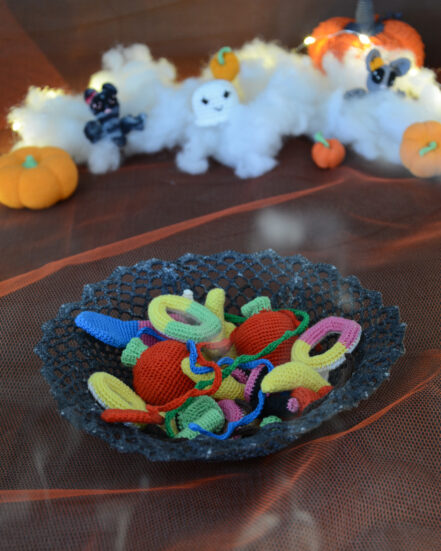 Bol en dentelle grise foncée rempli de bonbons colorés au crochet, posé sur un tissu orange, avec en arrière-plan des décorations d'Halloween crochetées : citrouilles, fantômes et chauve-souris sur un fond de nuages blancs.