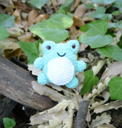 Petite grenouille au crochet en fil chenille vert d'eau avec un ventre blanc, posée au milieu de feuilles sèches et de lierre vert dans la nature.