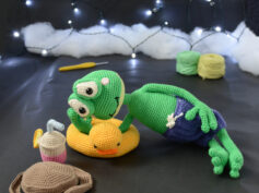Grenouille amigurumi verte allongée sur une bouée canard jaune, portant un short violet, avec un chapeau et une boisson crochetés posés à côté, sur fond sombre décoré de guirlande lumineuse.