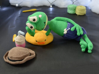 Jeannot la grenouille (grand) 14 Grenouille amigurumi verte allongée sur une bouée canard jaune, portant un short violet, avec un chapeau et une boisson crochetés posés à côté, sur fond sombre décoré de guirlande lumineuse.