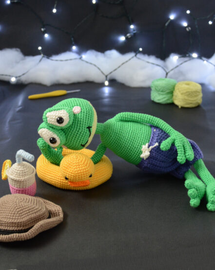 Grenouille amigurumi verte allongée sur une bouée canard jaune, portant un short violet, avec un chapeau et une boisson crochetés posés à côté, sur fond sombre décoré de guirlande lumineuse.