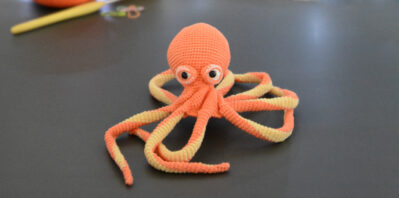 Un poulpe crocheté en corail et jaune pastel est posé comme dans un mouvement vers l'arrière avec sa tête sur un fond gris foncé avec en fond 2 pelotes de la même couleur que le poulpe, un crochet et des anneaux marqueurs. En arrière plan, il y a une guirlande lumineuse et un nuage de ouate.