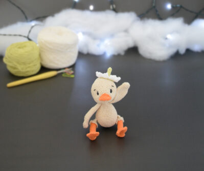 Amigurumi représentant un caneton crème qui fait coucou avec une aile. Il a un bec et des pattes orange, une fleur blanche sur la tête. Il est assis devant deux pelotes de coton, un crochet et une guirlande lumineuse en arrière-plan.