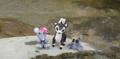 Trois amigurumis crochetés sont posés sur un rocher au bord d'une rivière. On voit une souris grise avec un nœud rose, un cerf marron et blanc et une chauve-souris grise avec un nœud orange. Derrière eux, de l'eau coule doucement. L'ambiance est calme et naturelle. La vue est prise légèrement au-dessus d'eux