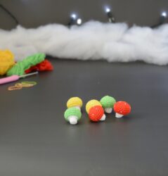 Six petits champignons crochetés en coton coloré. Leurs chapeaux sont rouge, jaune et vert. Ils sont posés sur un fond gris foncé. En arrière-plan, des pelotes et un crochet sont partiellement visibles, ainsi qu'un peu de rembourrage blanc et une guirlande lumineuse.