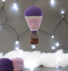 Montgolfière crochetée suspendue dans un décor gris avec des guirlandes lumineuses. Au sol, légèrement en arrière plan, pelotes de laine violette et lilas, crochet et nuages en ouate.