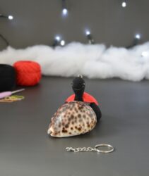 Une toute petite tortue crochetée en noir, avec une carapace rouge, posée devant un coquillage tacheté. Elle porte un anneau de porte-clés sur la tête. En arrière-plan : des pelotes rouge et noire, des anneaux marqueurs, un crochet, du rembourrage blanc et une guirlande lumineuse floue. À l'avant-plan se trouve la chaînette du porte-clés.